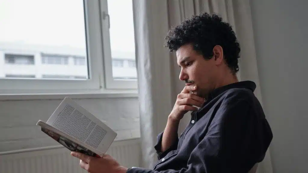 A man thoughtfully reading a book while sitting by a window.