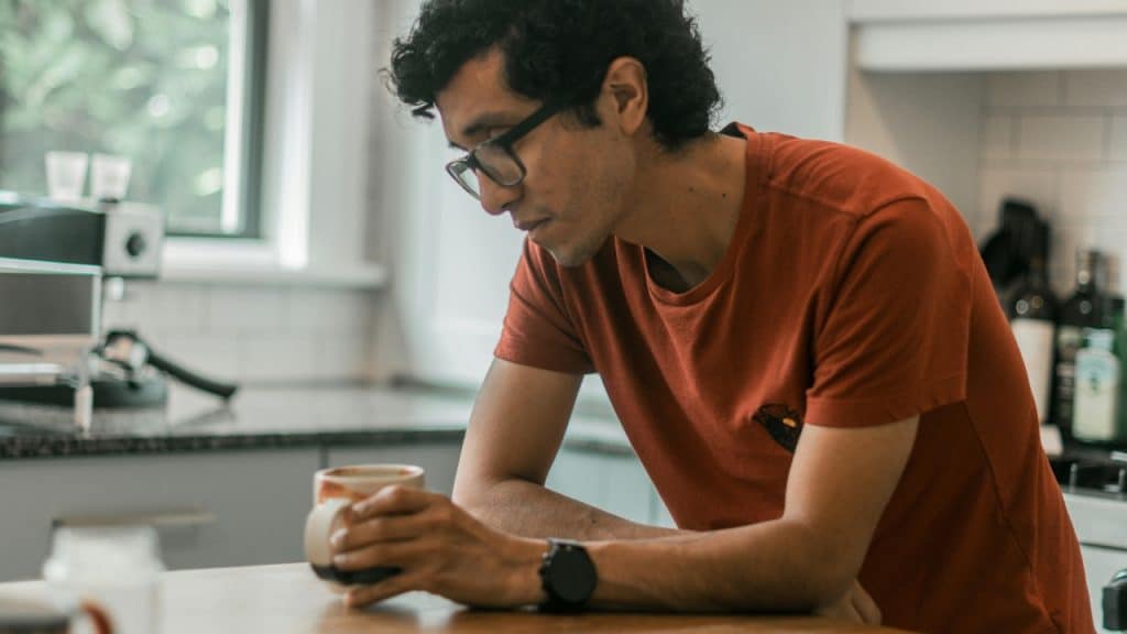 A man sits at a table in a kitchen, holding a mug and looking down.