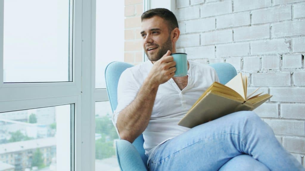 A smiling man sits in a blue armchair by a window, holding a book and mug.