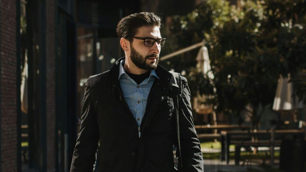 A bearded man with glasses wears a black jacket and blue shirt outdoors.