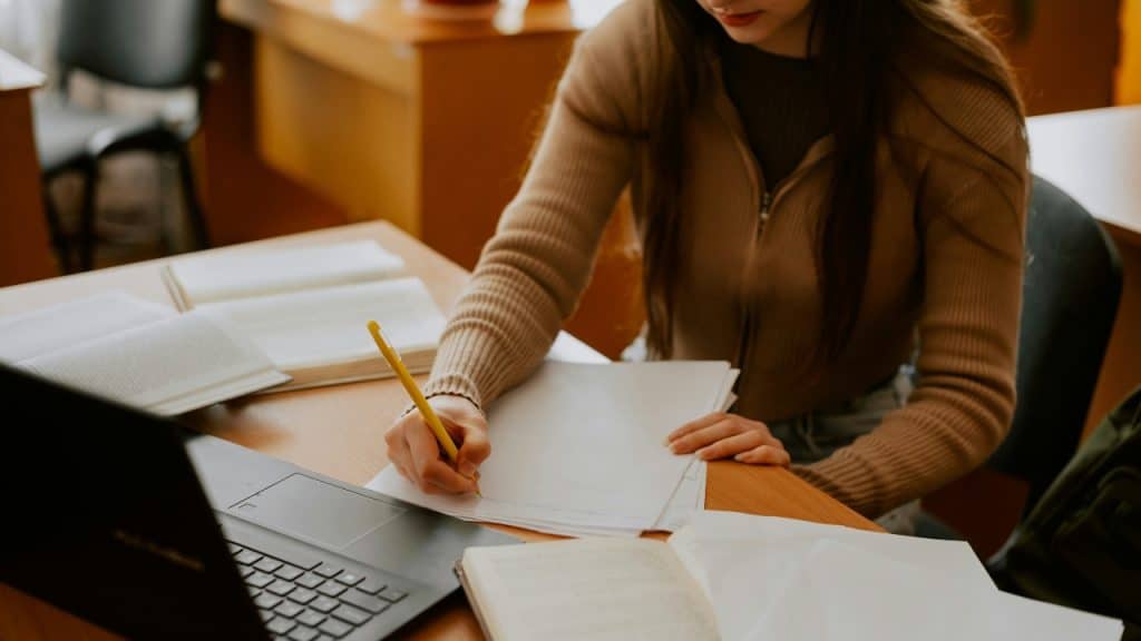 A person in a tan sweater writes on paper at a desk with a laptop.