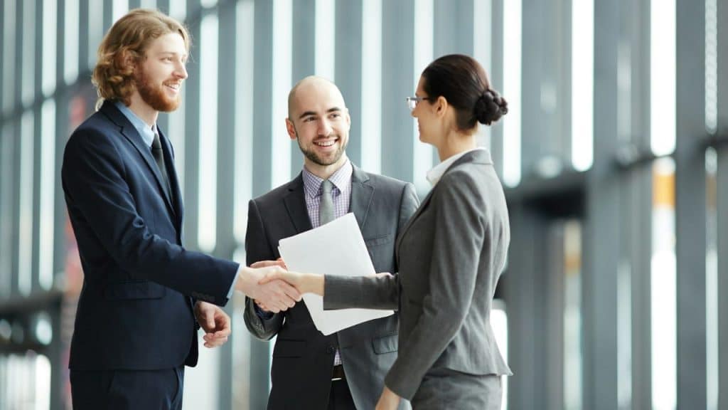 Three professionals in business attire shaking hands and smiling in a modern office hallway.