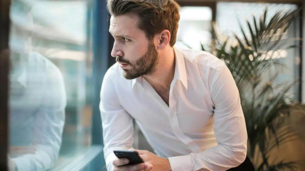 A man leaning by a window holding his phone and looking outside.