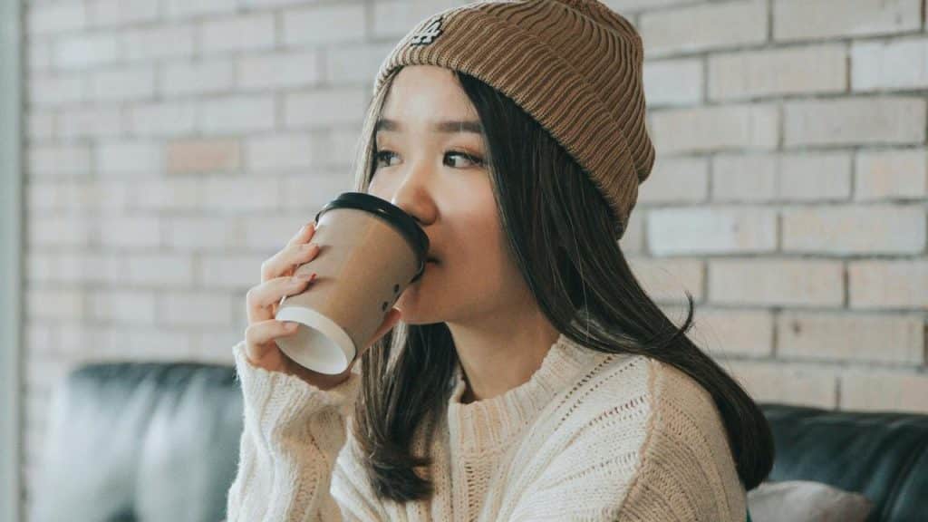 A woman wearing a knit beanie and sweater drinking from a takeaway coffee cup indoors.