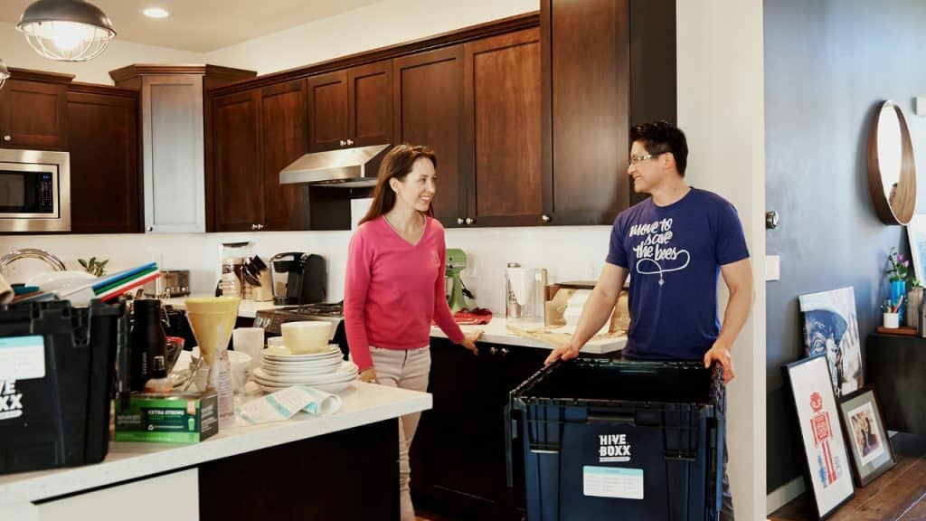 A man and woman talk in a kitchen near several large black plastic bins.