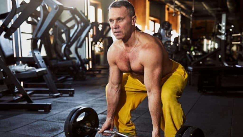 A shirtless man in yellow pants crouches while holding a barbell in a gym setting.