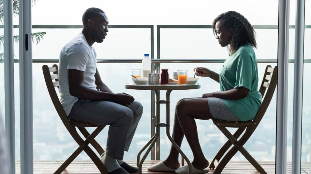 A man and woman sit facing each other at a small table on an outdoor balcony.