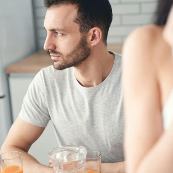 A man looks away in a kitchen, seemingly upset during a conversation with an out-of-focus woman.