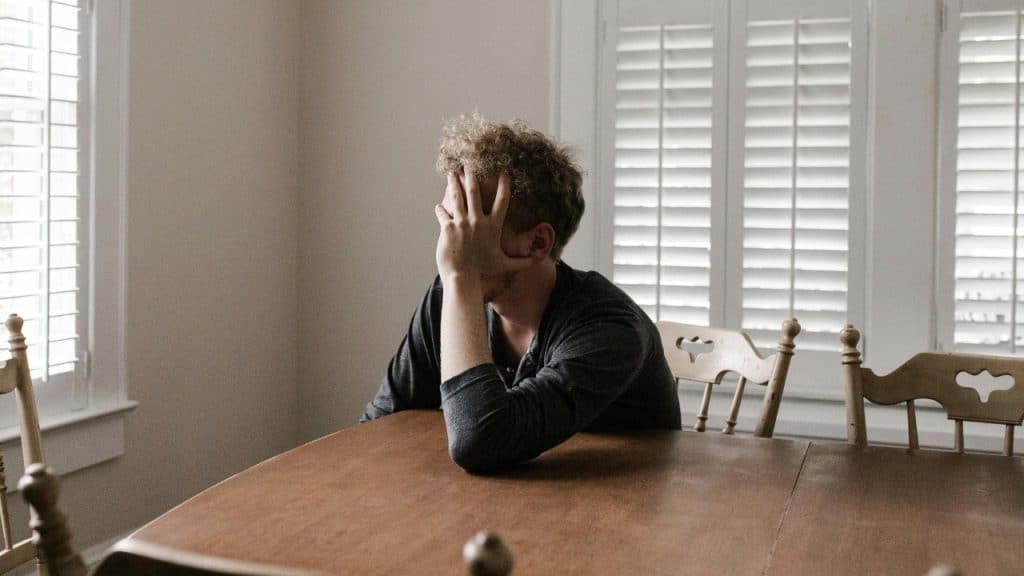 A person sits at a wooden dining table with their hand covering their face near windows.