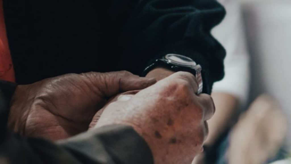 An elderly person's hands gently hold the wrist of another wearing a dark wristwatch.