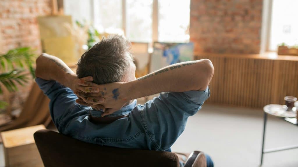 A person with paint-stained hands sits in a chair with their hands behind their head.