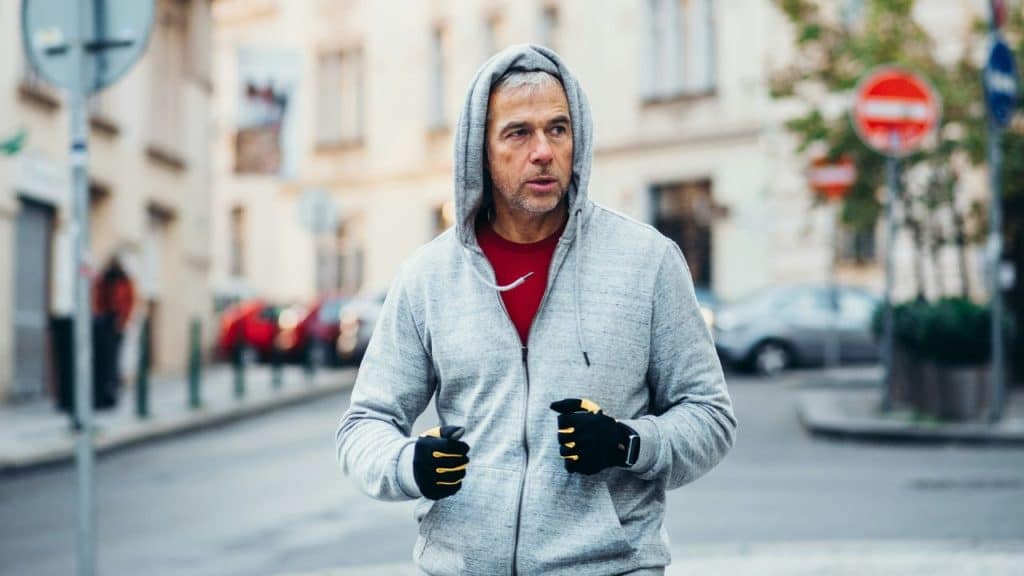 A man in a hooded sweatshirt and gloves jogging down a city street.