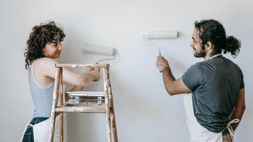 A man and woman stand on opposite sides of a wall, painting it white together.