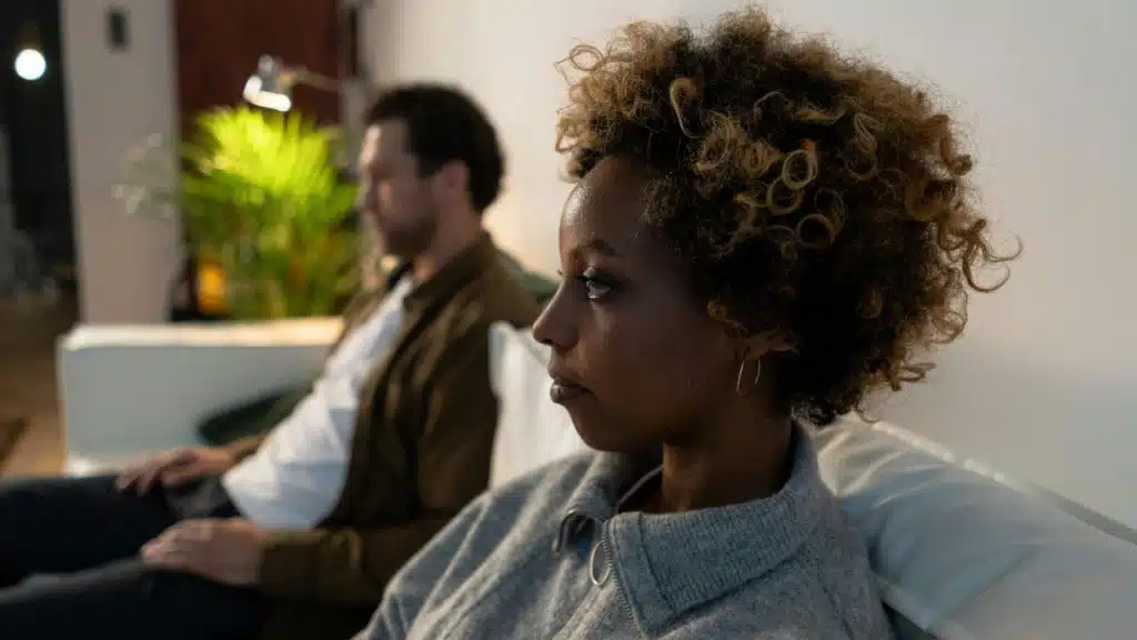 A woman in a grey sweater sits on a couch beside a blurred man.