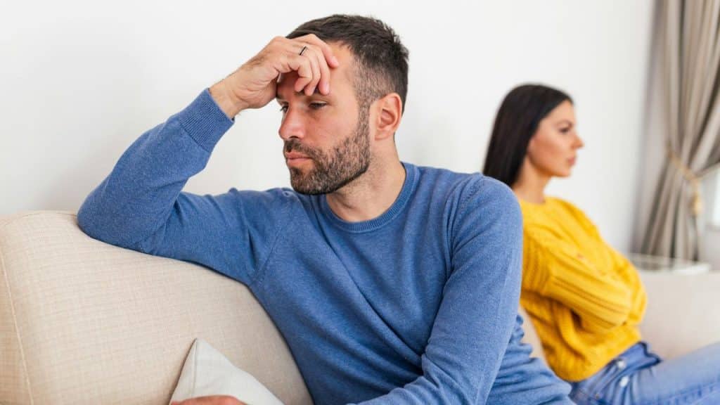 A man in a blue sweater touches his forehead while a woman sits behind him.