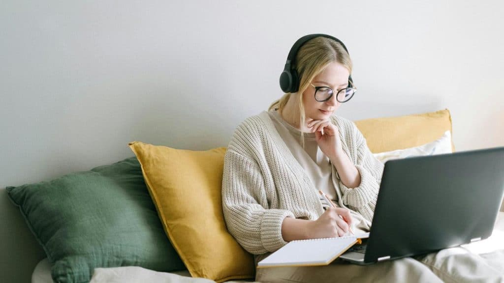 A woman wearing headphones taking notes while using a laptop.