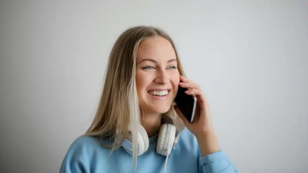 A smiling woman talking on a smartphone with headphones around her neck.