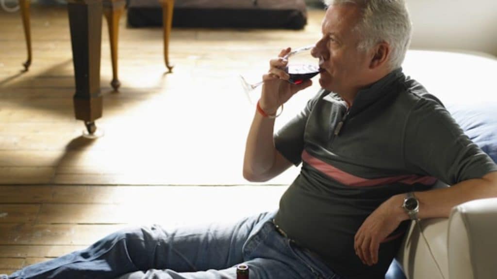 A man with gray hair sits on the floor, drinking from a wine glass.