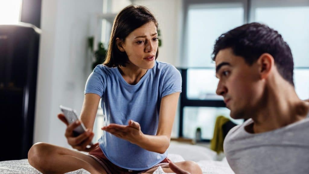 A woman holding a phone speaks to a man, gesturing while looking concerned.