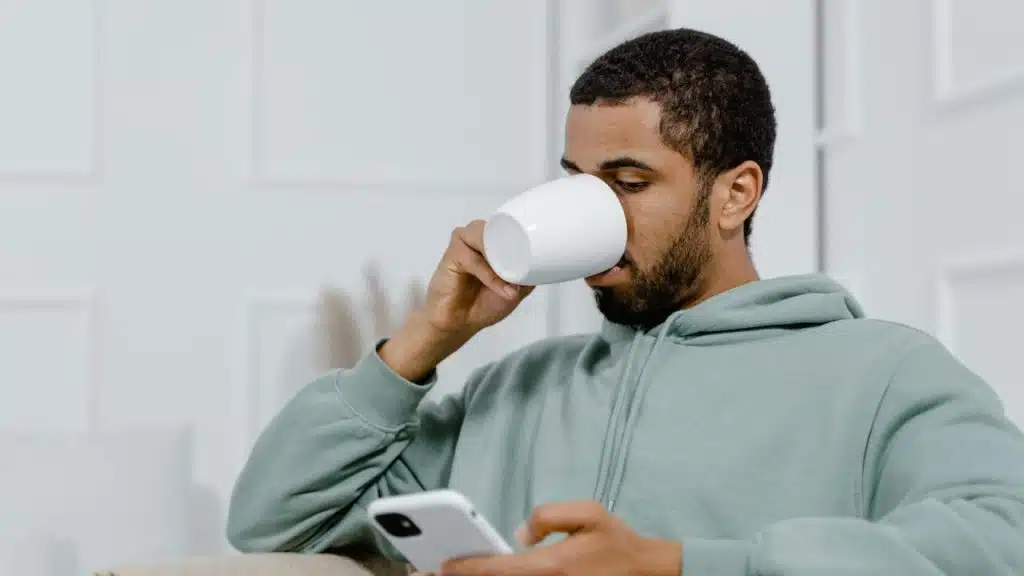 A man drinking from a mug while looking at his phone.