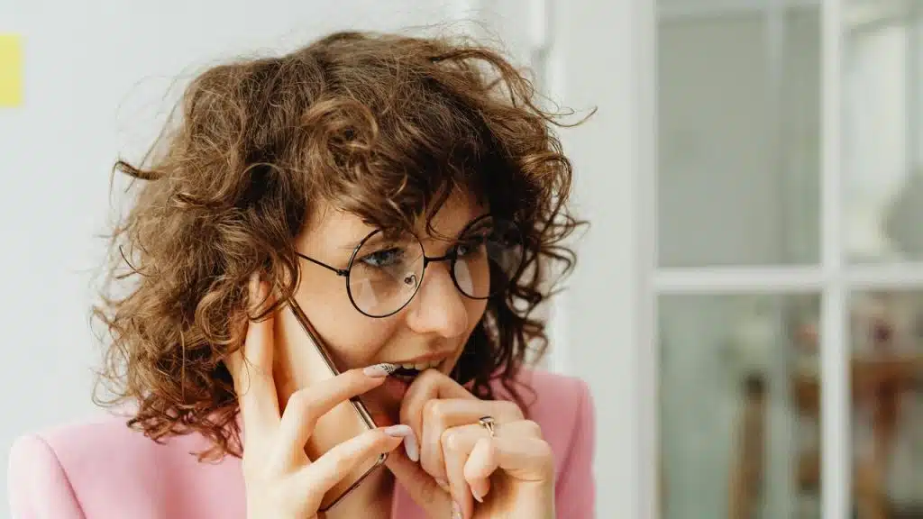 A worried woman with curly hair talks on the phone while biting her nails.