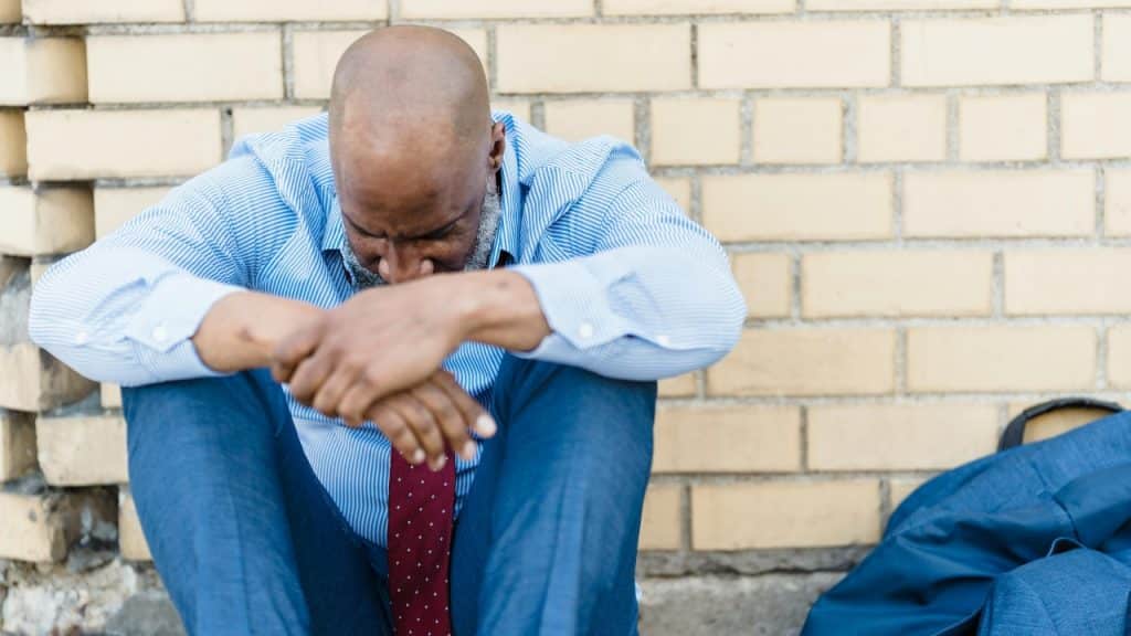 A bald man in a dress shirt sits on the ground, leaning against a brick wall.