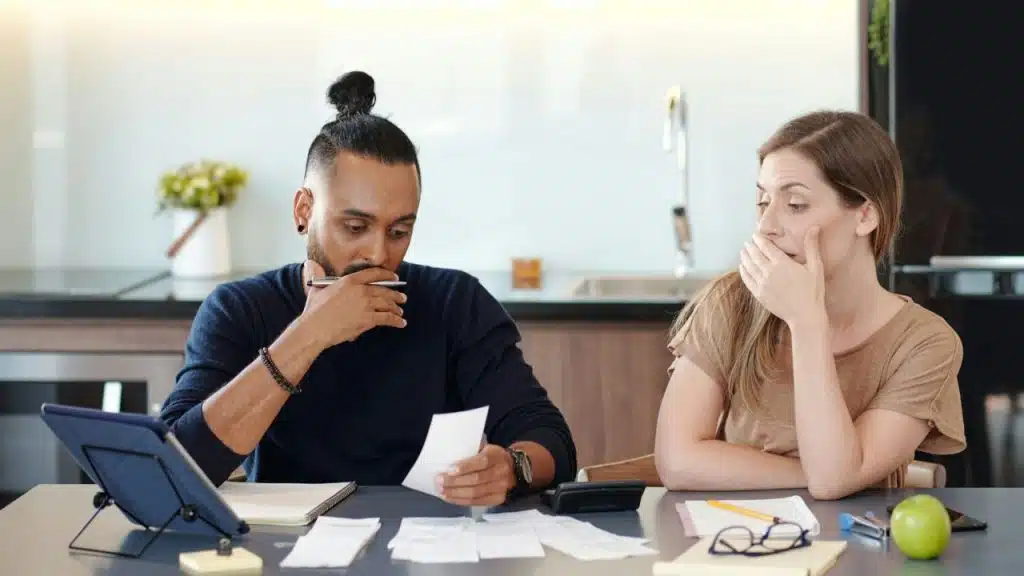 A couple sits at a table with receipts, looking worried while reviewing their finances.
