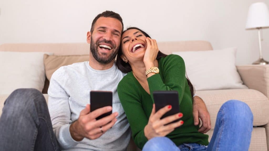 A man and woman sit on the floor laughing while holding their mobile phones.