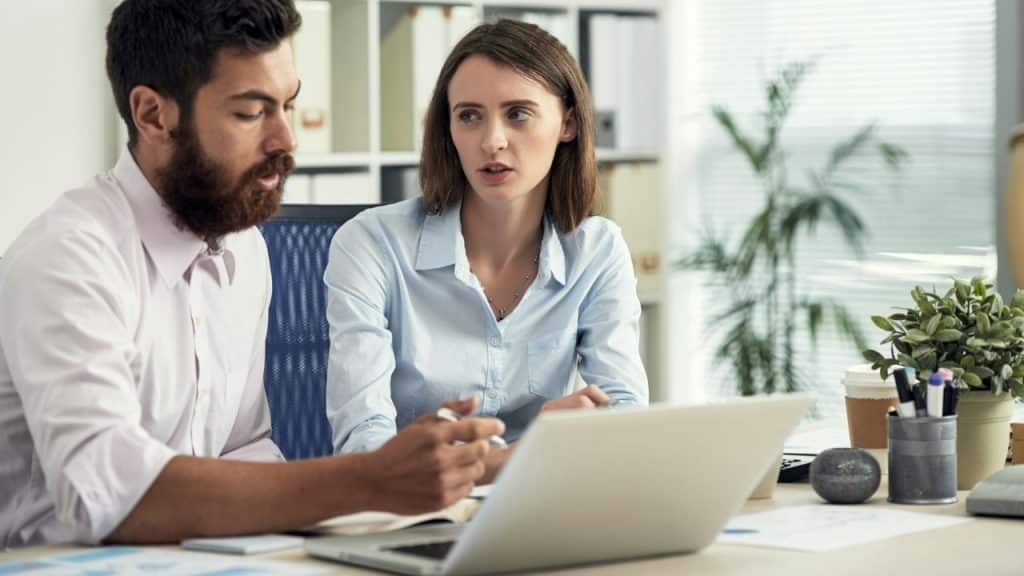 A man and a woman look at a laptop together while working in an office.