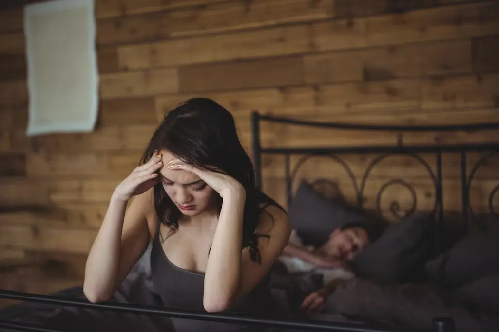 Upset woman sitting on a bed while man sleeping in background