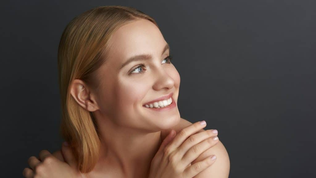 Close-up of a smiling woman with blonde hair looking upward against a dark grey background.