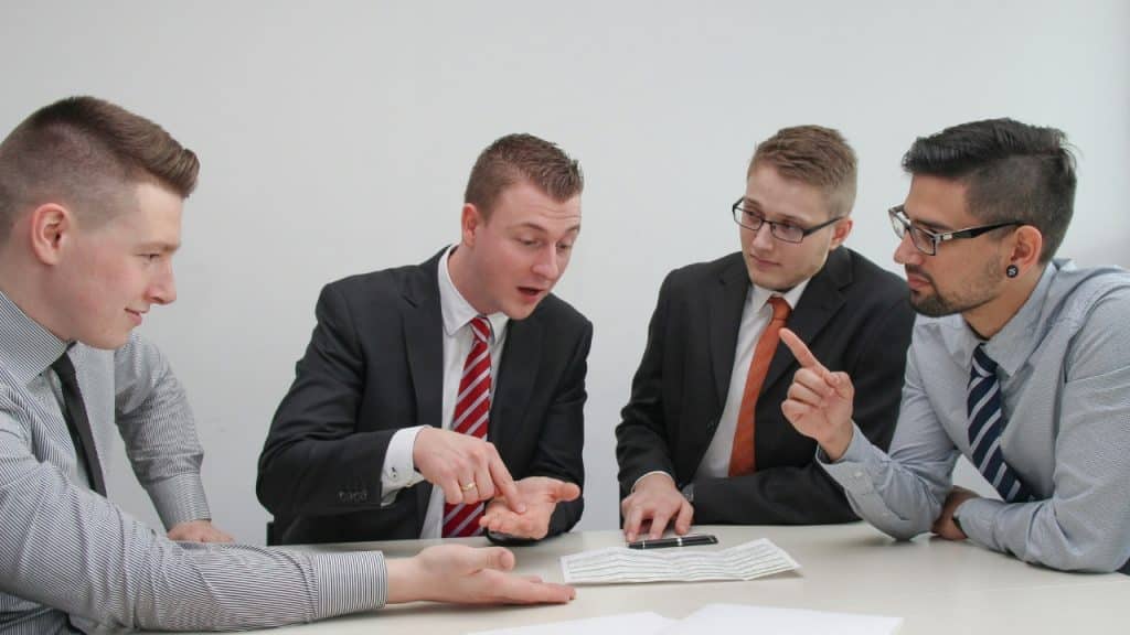 Four men in suits sit at a table with a document, gesturing and looking downward.