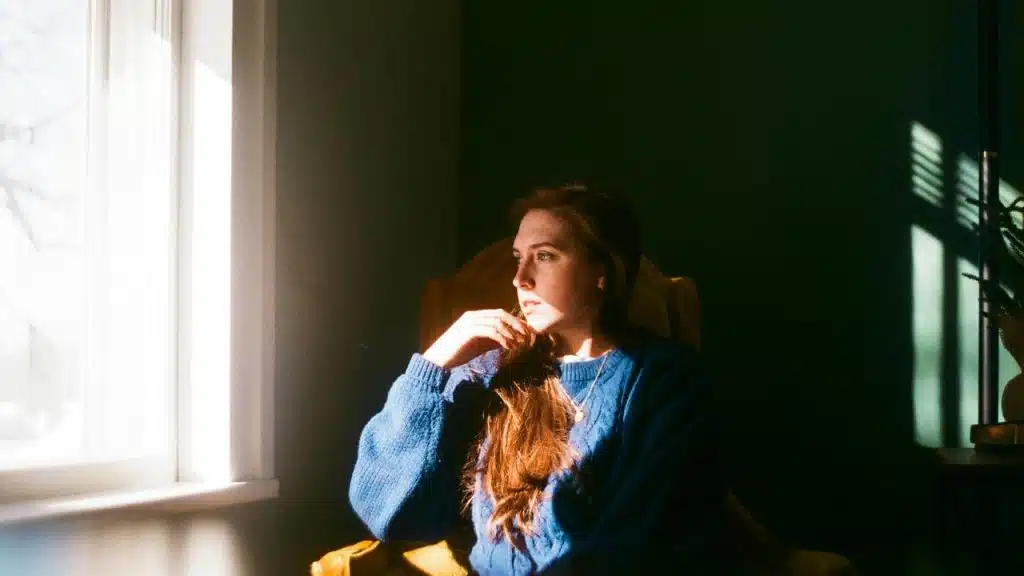 A woman sitting by a window looking outside in sunlight.