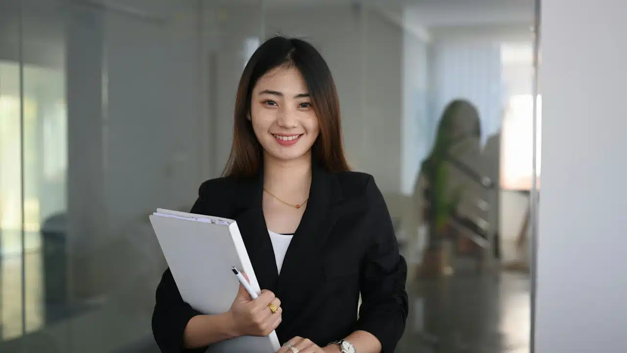 A smiling businesswoman in a black blazer stands in a modern office hallway holding a notebook and pen.