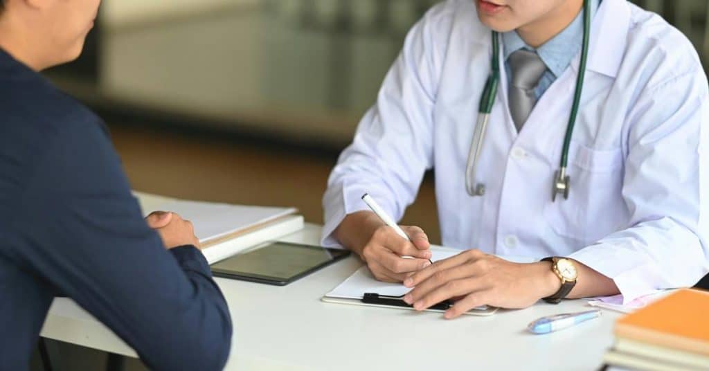 A doctor writing notes while consulting with a patient at a desk.