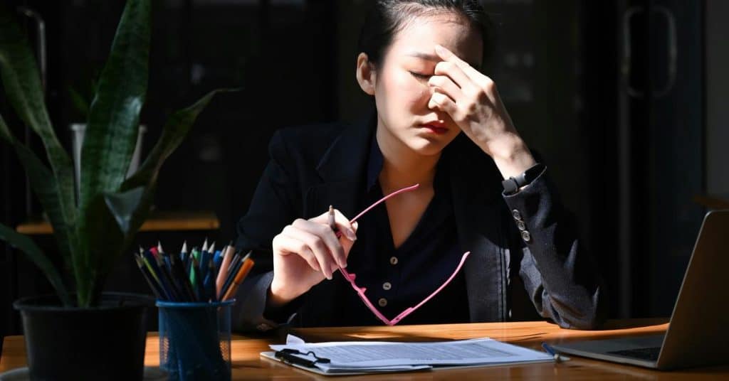 A woman at a desk holding glasses and rubbing her forehead, appearing stressed.