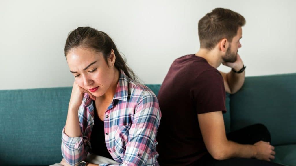 A woman and man sit back-to-back on a blue sofa with their heads down.