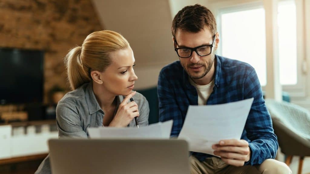 A man and woman look seriously at paperwork while sitting in front of a laptop.