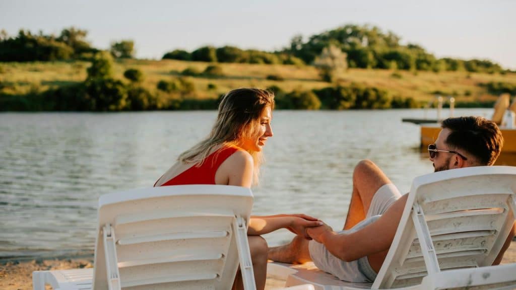 A couple relaxing on lounge chairs by a lake, holding hands and talking.
