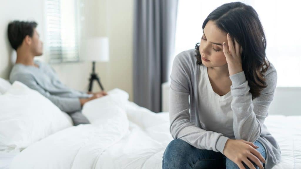 A woman sits on a bed holding her head while a man sits blurred in the background.