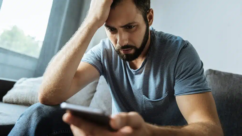 A bearded man sits on a couch holding his head while looking at a smartphone.