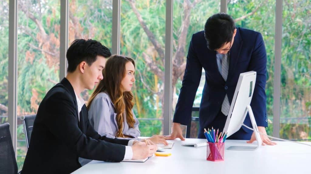 Three people in business attire work at a white desk in a bright office.