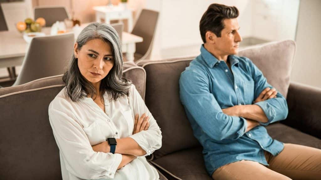 A mature older couple sitting on a couch with arms crossed and refusing to look at each other. 