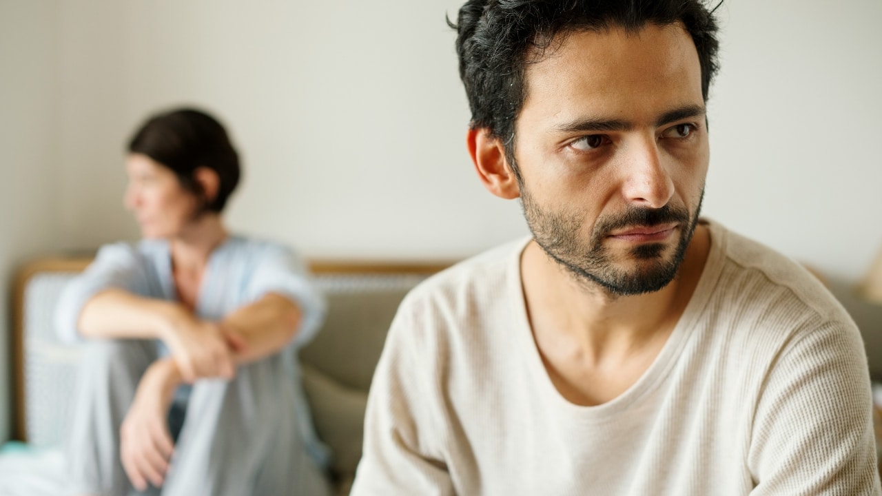 A man in a white shirt looks away while a woman sits blurred behind him.