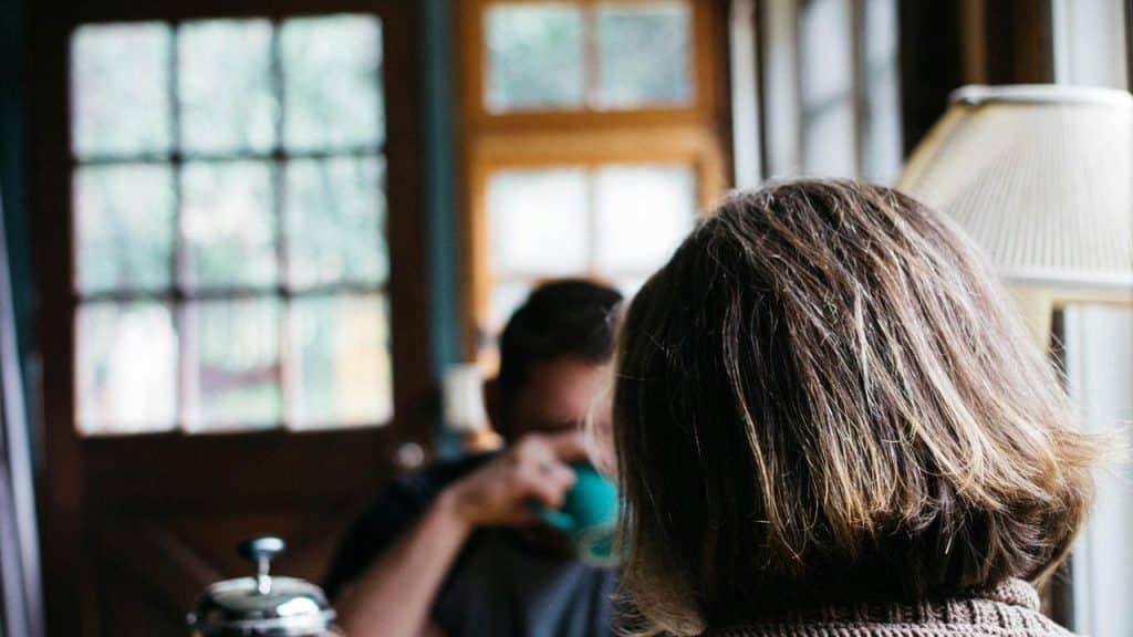 A person sitting indoors with another person blurred in the background.