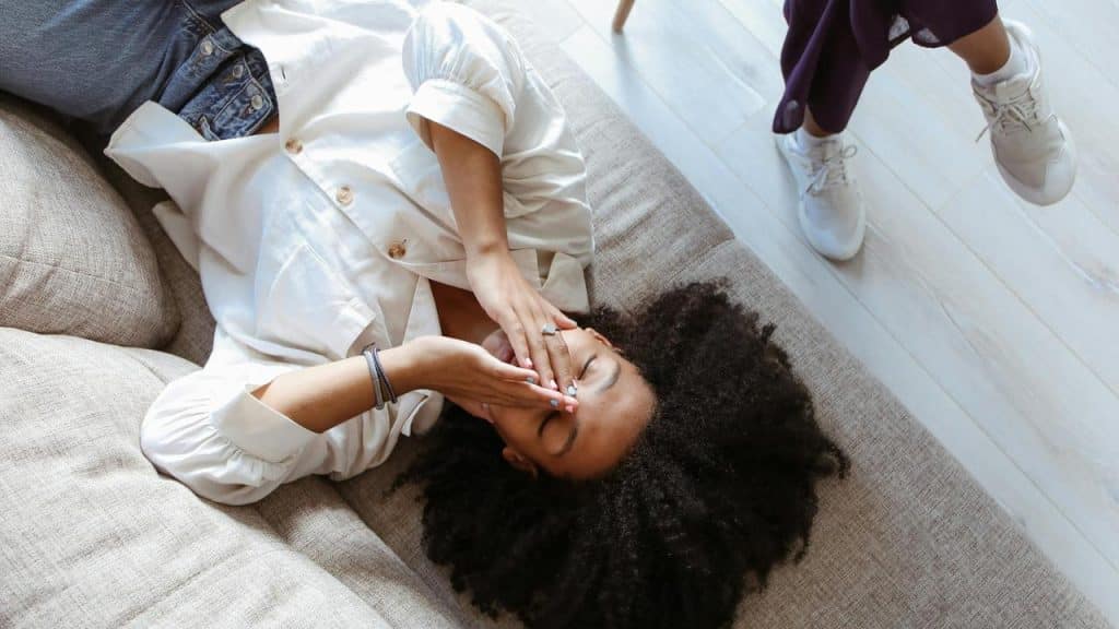A woman lies on a couch covering her face while another person stands nearby.