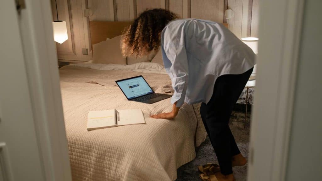 A woman leaning over a bed with a laptop and notebook in a softly lit bedroom.