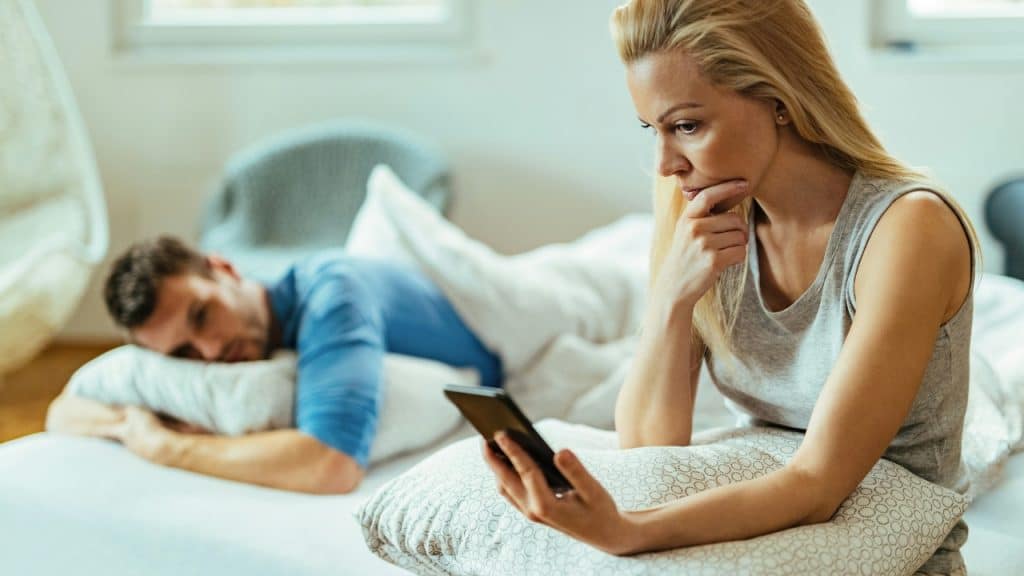 A woman sits on a bed looking at a phone while a man lies behind her.