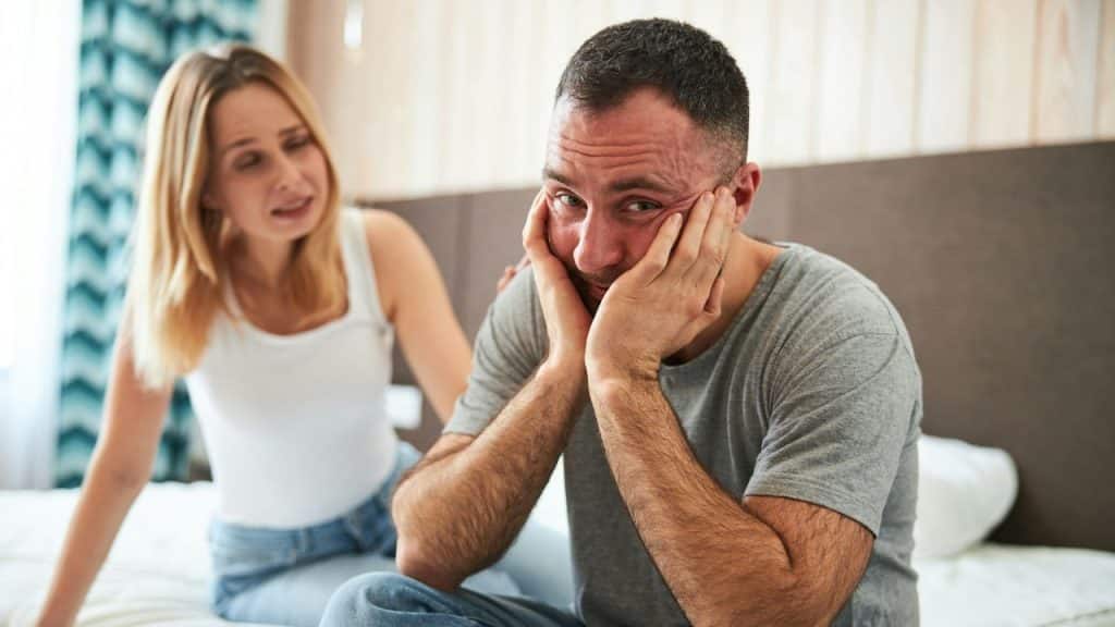 A man holding his face in his hands sits on a bed next to a woman.