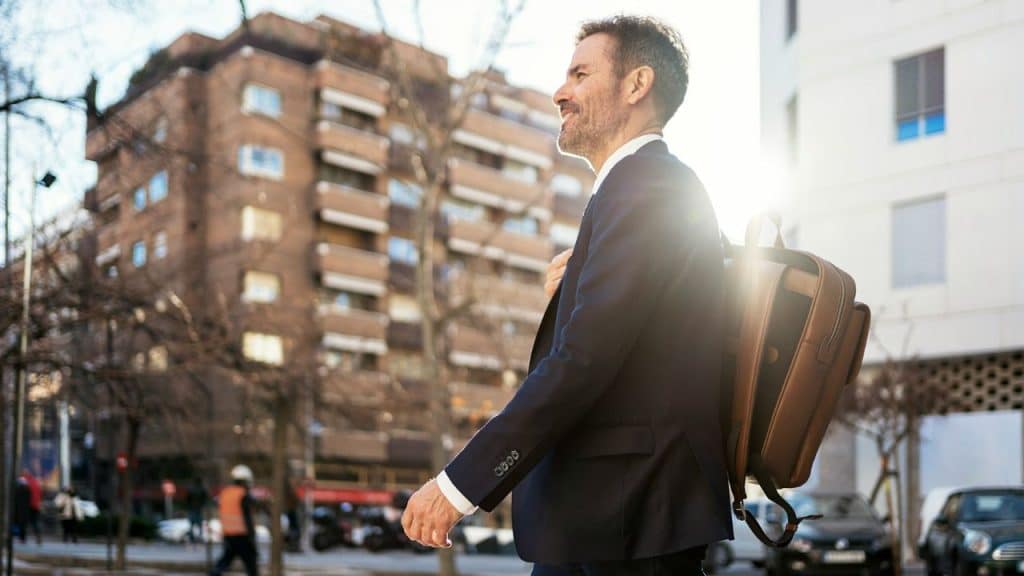 A man in a navy suit and leather backpack walks down a city street in bright sunlight.